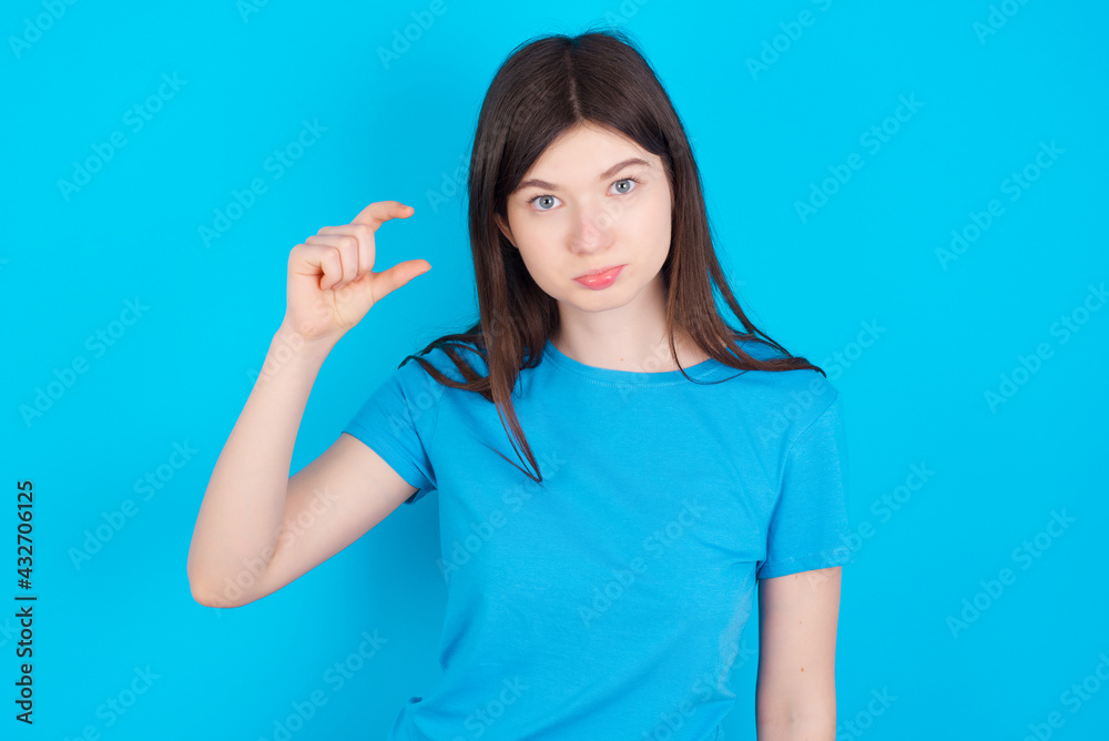 young beautiful Caucasian woman wearing blue T-shirt over blue wall purses lip and gestures with hand, shows something very little. young beautiful Caucasian woman wearing blue T-shirt over blue wall purses lip and gestures with hand, shows something very little.