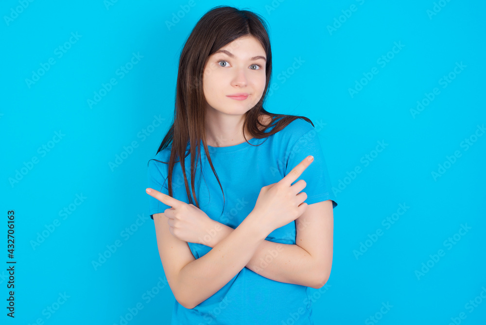 young beautiful Caucasian woman wearing blue T-shirt over blue wall crosses arms and points at different sides hesitates between two items or variants. Needs help with decision young beautiful Caucasian woman wearing blue T-shirt over blue wall crosses arms and points at different sides hesitates between two items or variants. Needs help with decision