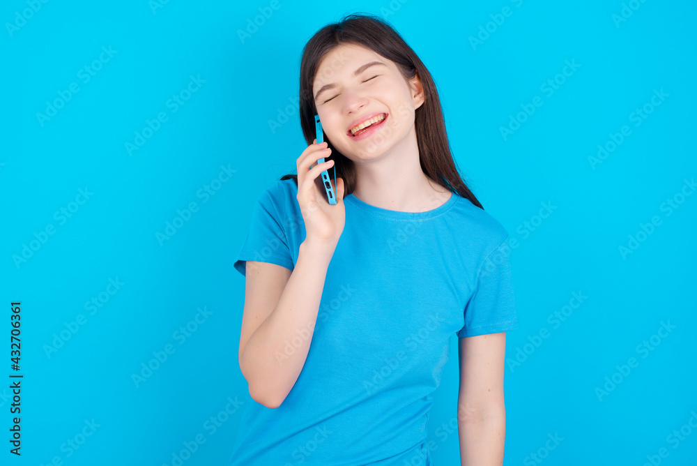 Funny young beautiful Caucasian woman wearing blue T-shirt over blue wall laughs happily, has phone conversation, being amused by friend, closes eyes. Funny young beautiful Caucasian woman wearing blue T-shirt over blue wall laughs happily, has phone conversation, being amused by friend, closes eyes.