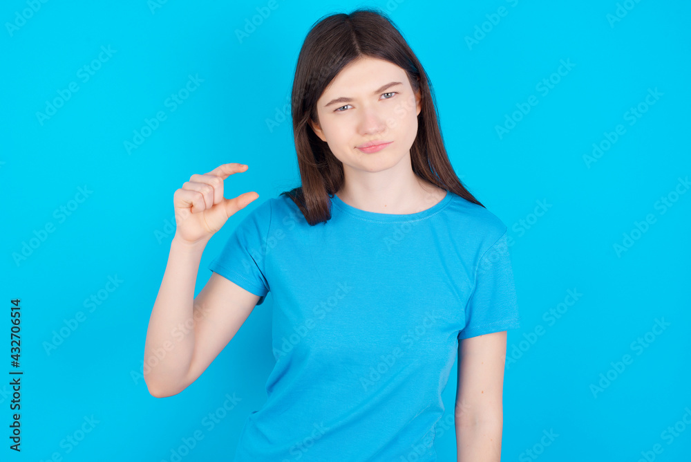 Displeased young beautiful Caucasian woman wearing blue T-shirt over blue wall shapes little hand sign demonstrates something not very big. Body language concept. Displeased young beautiful Caucasian woman wearing blue T-shirt over blue wall shapes little hand sign demonstrates something not very big. Body language concept.