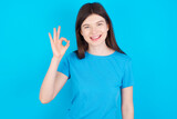 young beautiful Caucasian woman wearing blue T-shirt over blue wall hold hand arm okey symbol toothy approve advising novelty news