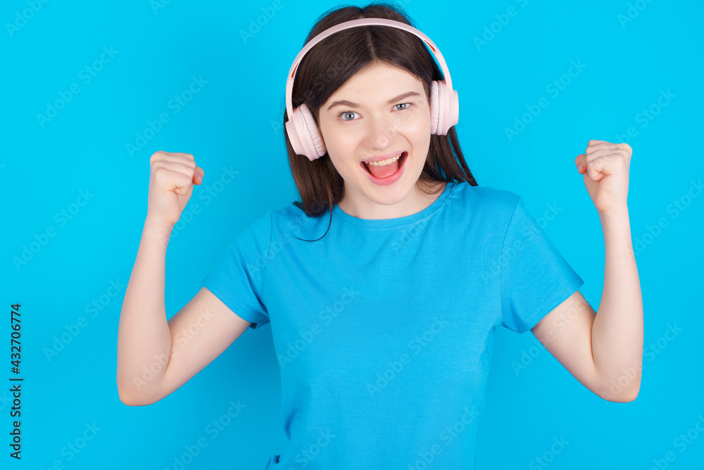 Emotional young beautiful Caucasian woman exclaims loudly feels like winner raises clenched fists keeps mouth opened wears stereo headphones on ears makes yes gesture, listens favourite music Emotional young beautiful Caucasian woman exclaims loudly feels like winner raises clenched fists keeps mouth opened wears stereo headphones on ears makes yes gesture, listens favourite music