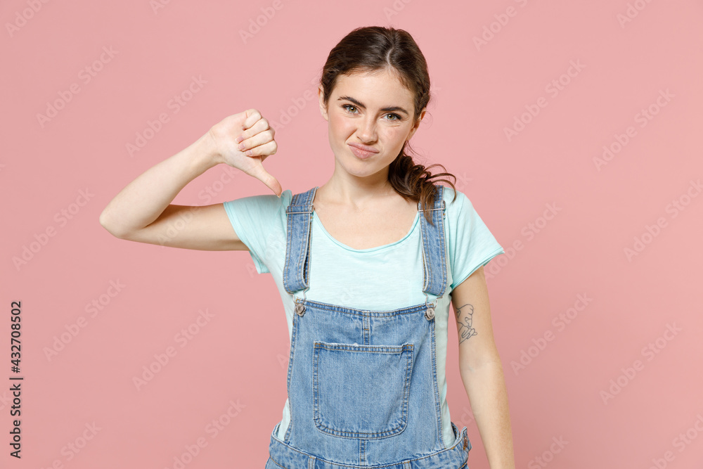 Young displeased sad disappointed caucasian woman 20s wearing denim clothes blue t-shirt showing thumb down gesture isolated on pastel pink color background studio portrait. People lifestyle concept Young displeased sad disappointed caucasian woman 20s wearing denim clothes blue t-shirt showing thumb down gesture isolated on pastel pink color background studio portrait. People lifestyle concept