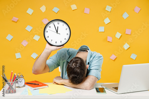 Young tired exhausted sad employee business man wearing shirt sit work white office desk with pc laptop hold clock putting hand on desk sleeping isolated on yellow color background studio portrait