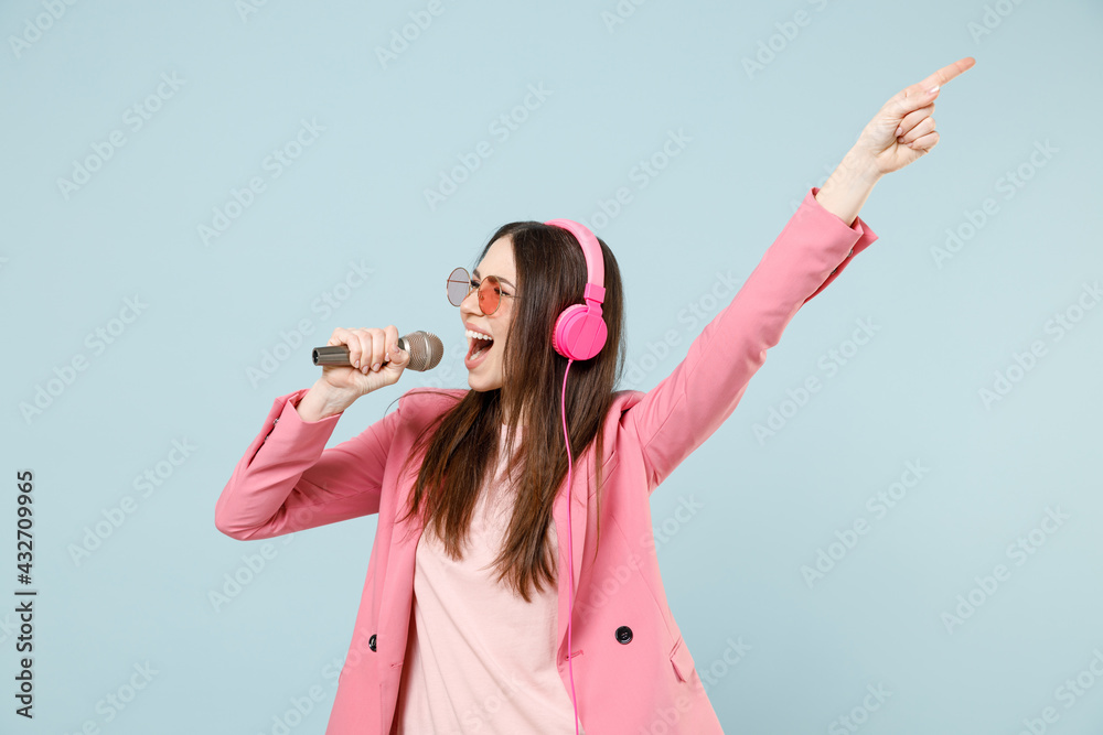 Young woman 20s wear pastel pink clothes glasses headphones sing song in microphone at karaoke club with outstretched hand isolated on blue color background studio People lifestyle technology concept.