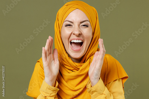 Close up young expressive arabian asian muslim woman 20s wearing abaya hijab yellow clothes scream news with hands near mouth isolated on olive green background. People uae islam religious concept
