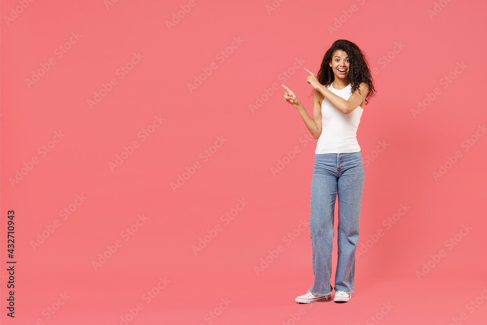 Full length young fun excited happy positive african american woman 20s wear casual white tank shirt walk go point index finger aside on copy space area mock up workspace isolated on pink background Full length young fun excited happy positive african american woman 20s wear casual white tank shirt walk go point index finger aside on copy space area mock up workspace isolated on pink background