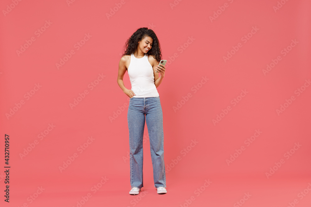 Full length young fun smiling happy satisfied positive african american woman in casual white tank shirt hold in hand mobile cell phone chat online browsing walking going isolated on pink background