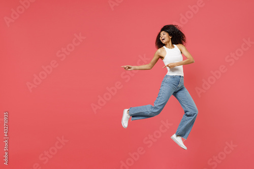 Fototapeta Naklejka Na Ścianę i Meble -  Full length young fun happy positive african american woman in casual white tank shirt jump high point index fingers aside on workspace area mock up isolated on pink color background studio portrait.