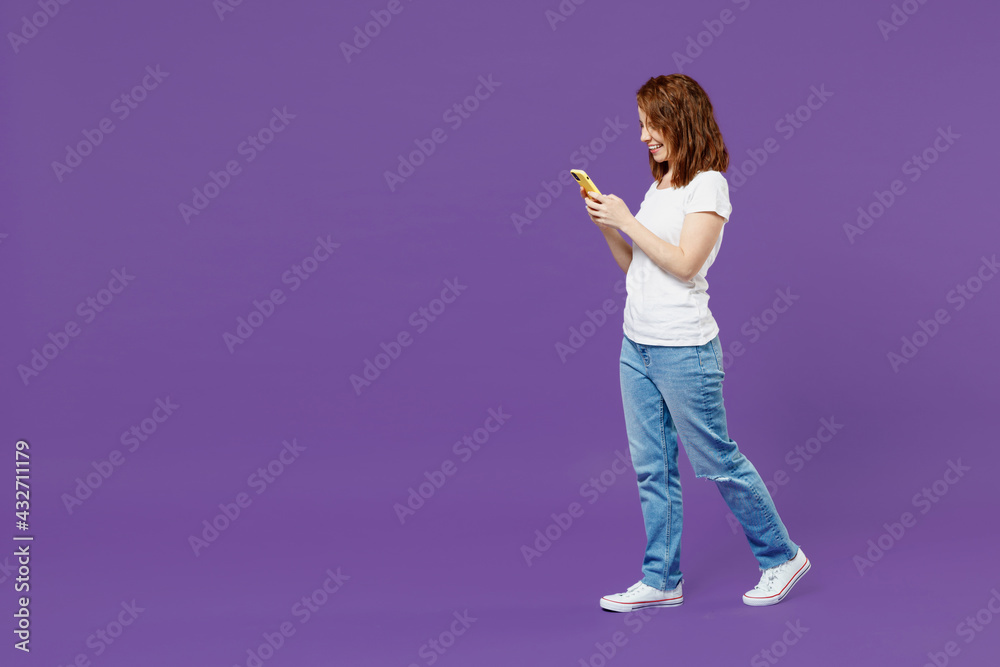 Full length side view young fun happy satisfied redhead woman in white basic casual t-shirt hold using mobile cell phone chat online browsing search isolated on dark violet background studio portrait