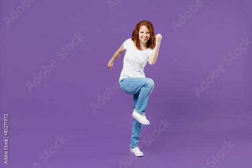 Full length young fun overjoyed redhead caucasian woman 20s wearing white basic casual t-shirt jeans do winner gesture clench fist with raised up leg isolated on dark violet color background studio