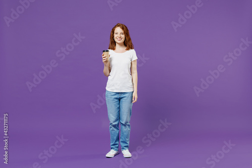 Full length young fun happy satisfied redhead caucasian woman 20s wearing white basic casual t-shirt hold paper cup of coffee drink tea look camera isolated on dark violet background studio portrait