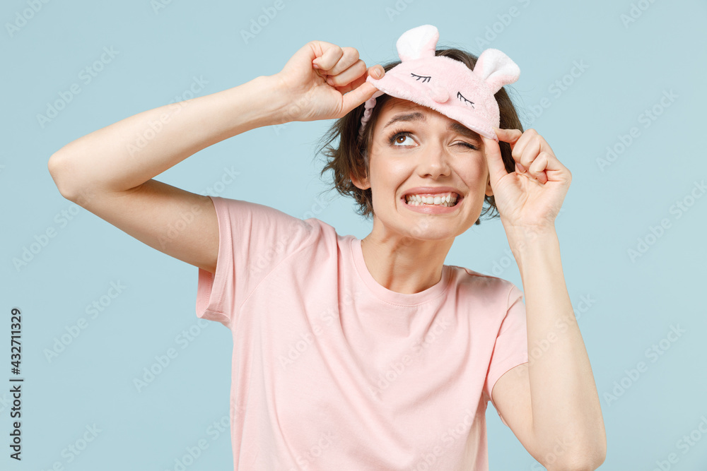 Young fun confused caucasian woman wear pajamas jam sleep rest relaxing at home took off blindfold eye mask isolated on pastel blue color background studio portrait. Good mood night bedtime concept. Young fun confused caucasian woman wear pajamas jam sleep rest relaxing at home took off blindfold eye mask isolated on pastel blue color background studio portrait. Good mood night bedtime concept.