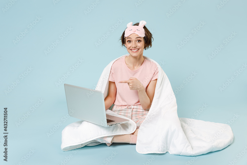 Young woman in pajamas jam sleep eye mask rest at home sit wrap cover under blanket duvet point index finger on laptop pc computer isolated on pastel blue background Good mood night bedtime concept. Young woman in pajamas jam sleep eye mask rest at home sit wrap cover under blanket duvet point index finger on laptop pc computer isolated on pastel blue background Good mood night bedtime concept.