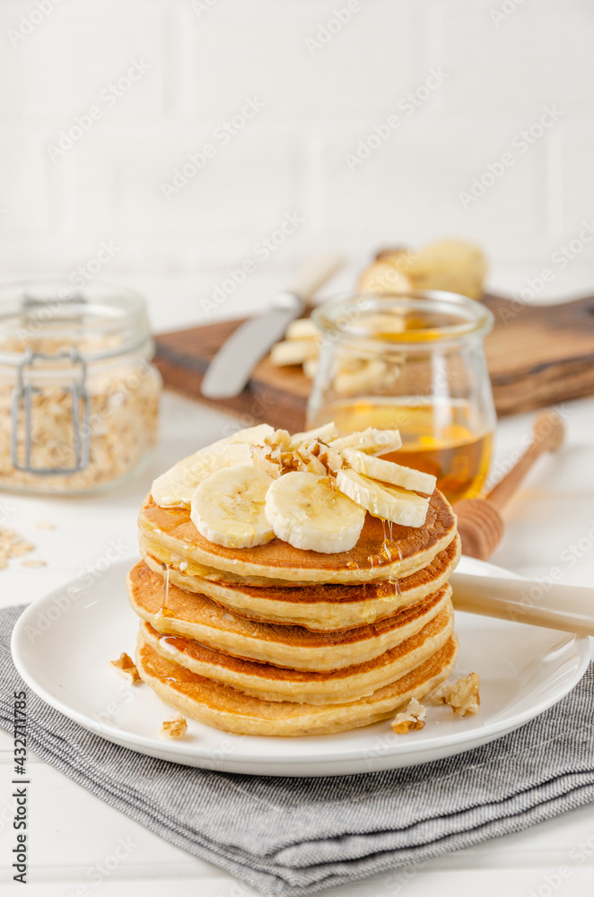 A stack of oatmeal banana pancakes with slices of fresh bananas, walnuts and honey on top with cup of tea on a white wooden background. A healthy breakfast. Copy space. A stack of oatmeal banana pancakes with slices of fresh bananas, walnuts and honey on top with cup of tea on a white wooden background. A healthy breakfast. Copy space.