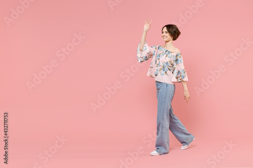 Full length young smiling fun woman 20s with short hairdo wear trendy stylish blouse walking go waving hand hello gesture isolated on pastel pink background studio portrait. People lifestyle concept