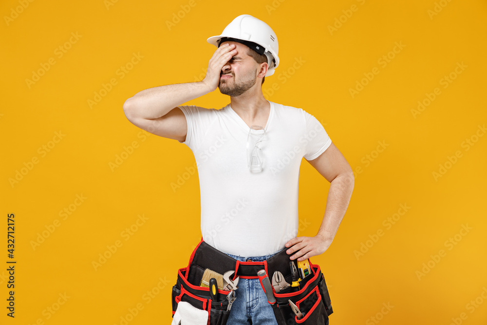 Young employee handyman man in protective helmet hardhat put hand face facepalm epic fail gesture isolated on yellow background. Instruments accessories renovation apartment room. Repair home concept. Young employee handyman man in protective helmet hardhat put hand face facepalm epic fail gesture isolated on yellow background. Instruments accessories renovation apartment room. Repair home concept.