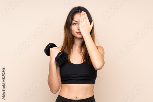 Young sport woman making weightlifting isolated on beige background covering a eye by hand