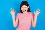 Emotive young beautiful Caucasian woman wearing pink T-shirt over blue wall laughs loudly, hears funny joke or story, raises palms with satisfaction, being overjoyed amused by friend