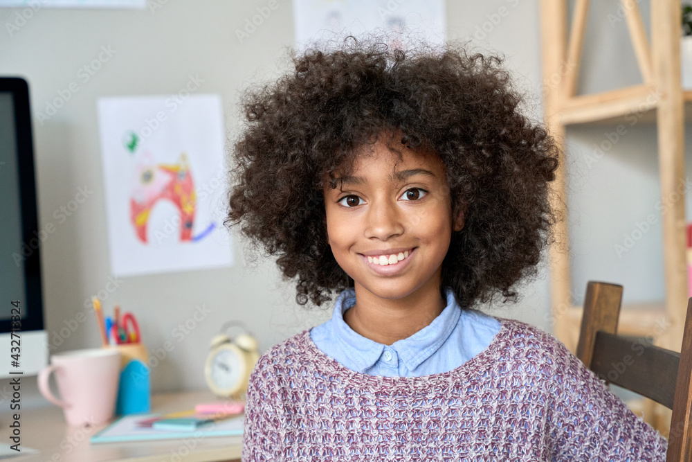 Fototapeta premium Happy cute smiling African American black preteen girl with curly afro hairstyle posing indoors in room looking at camera. Headshot closeup portrait of mixed race model.