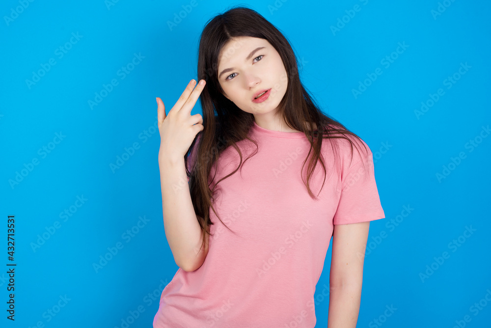 Unhappy young beautiful Caucasian woman wearing pink T-shirt over blue wall makes suicide gesture and imitates gun with hand, curves lips, keeps two fingers on temple Unhappy young beautiful Caucasian woman wearing pink T-shirt over blue wall makes suicide gesture and imitates gun with hand, curves lips, keeps two fingers on temple