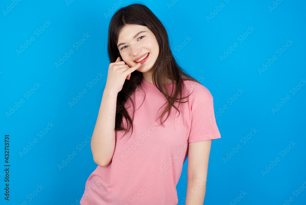 young beautiful Caucasian woman wearing pink T-shirt over blue wall smiling doing phone gesture with hand and fingers like talking on the telephone. Communicating concepts. young beautiful Caucasian woman wearing pink T-shirt over blue wall smiling doing phone gesture with hand and fingers like talking on the telephone. Communicating concepts.