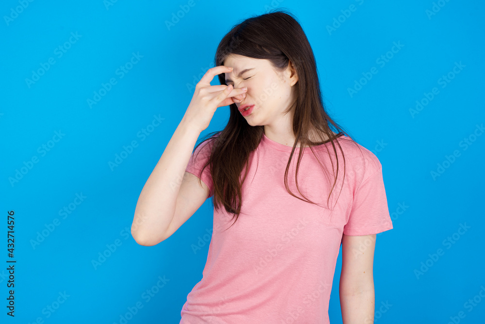 young beautiful Caucasian woman wearing pink T-shirt over blue wall smelling something stinky and disgusting, intolerable smell, holding breath with fingers on nose. Bad smell young beautiful Caucasian woman wearing pink T-shirt over blue wall smelling something stinky and disgusting, intolerable smell, holding breath with fingers on nose. Bad smell