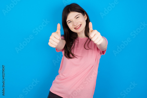 young beautiful Caucasian woman wearing pink T-shirt over blue wall approving doing positive gesture with hand, thumbs up smiling and happy for success. Winner gesture.