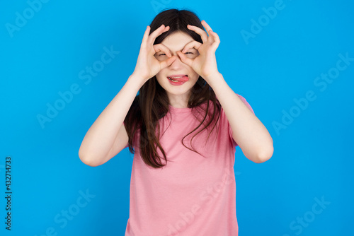 young beautiful Caucasian woman wearing pink T-shirt over blue wall doing ok gesture like binoculars sticking tongue out, eyes looking through fingers. Crazy expression.