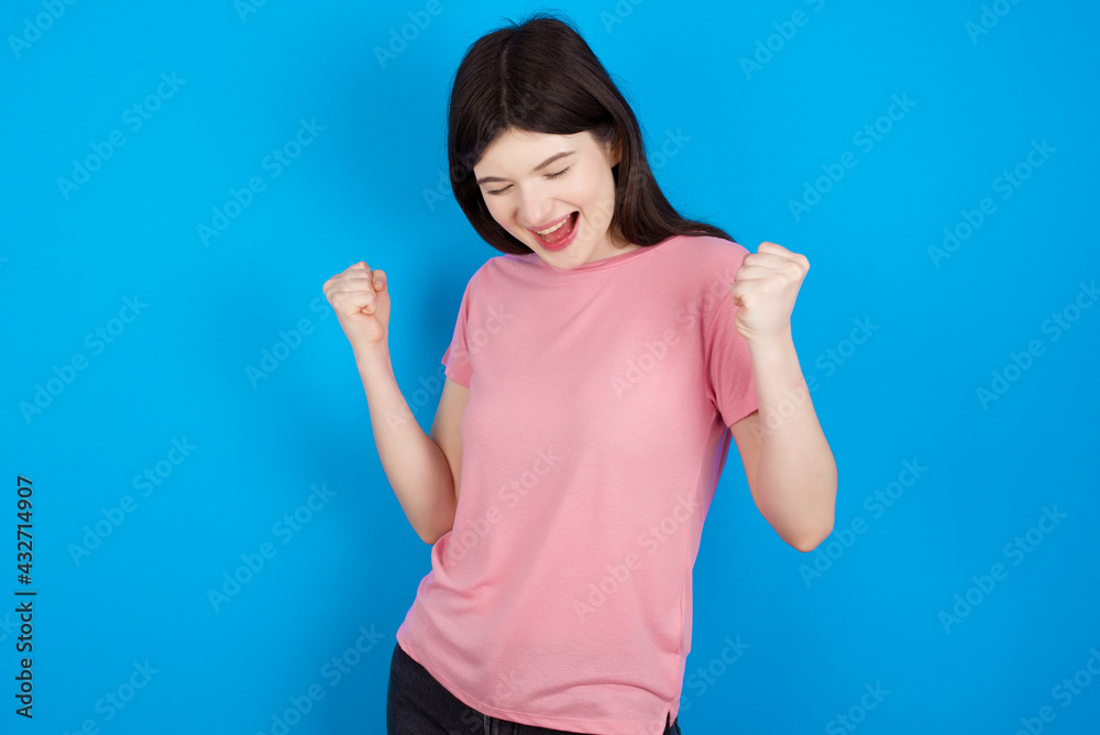 young beautiful Caucasian woman wearing pink T-shirt over blue wall very happy and excited doing winner gesture with arms raised, smiling and screaming for success. Celebration concept. young beautiful Caucasian woman wearing pink T-shirt over blue wall very happy and excited doing winner gesture with arms raised, smiling and screaming for success. Celebration concept.
