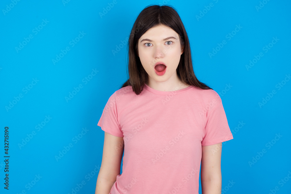 Obraz premium Oh my God. Surprised young beautiful Caucasian woman wearing pink T-shirt over blue wall stares at camera with shocked expression exclaims with unexpectedness,