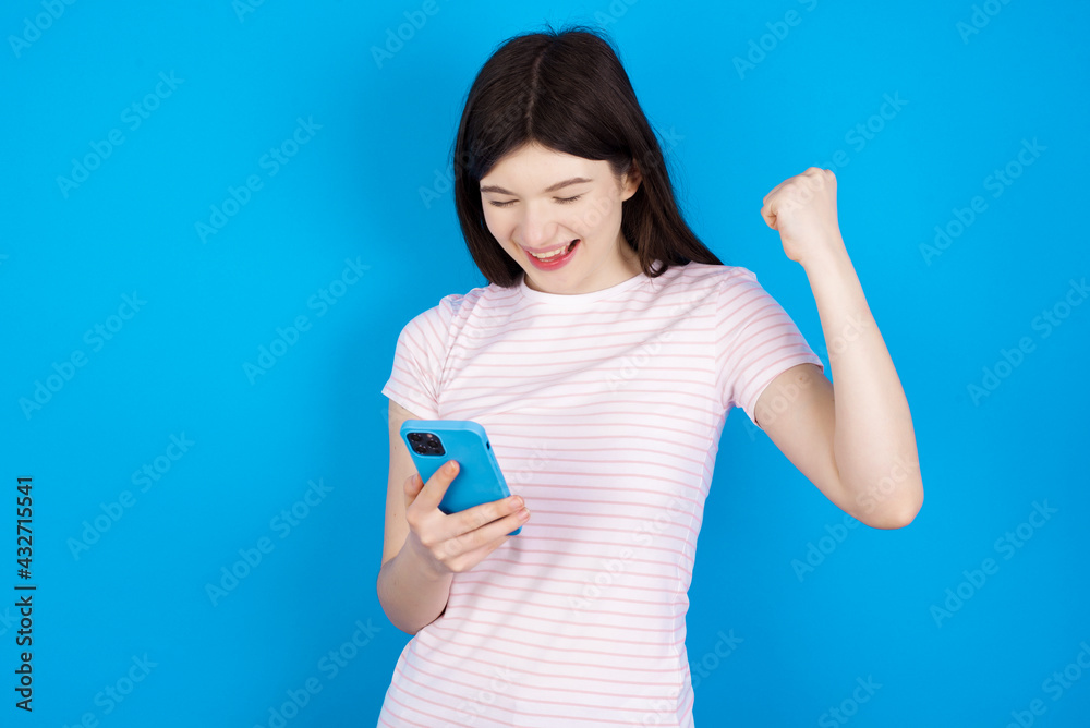 young beautiful Caucasian woman wearing stripped T-shirt over blue wall holding in hands cell and rising his fist up being excited after reading good news.