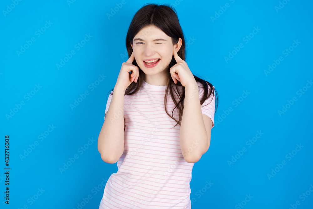 Happy young beautiful Caucasian woman wearing stripped T-shirt over blue wall ignores loud music and plugs ears with fingers asks to turn off sound Happy young beautiful Caucasian woman wearing stripped T-shirt over blue wall ignores loud music and plugs ears with fingers asks to turn off sound