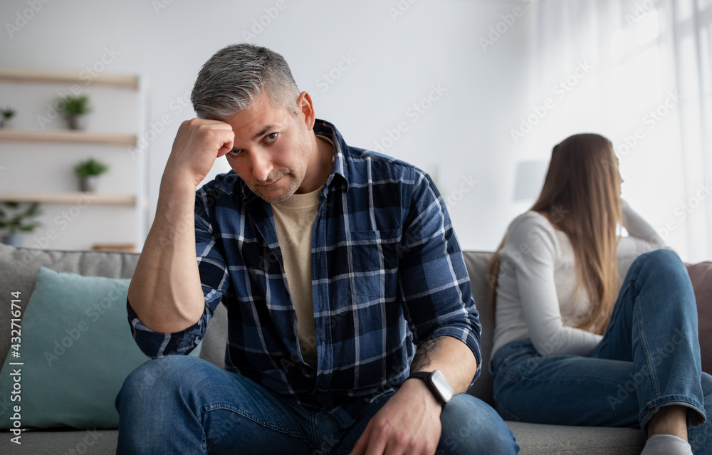 Upset mature man and his wife feeling offended after family fight, sitting on opposite ends of sofa at home, copy space