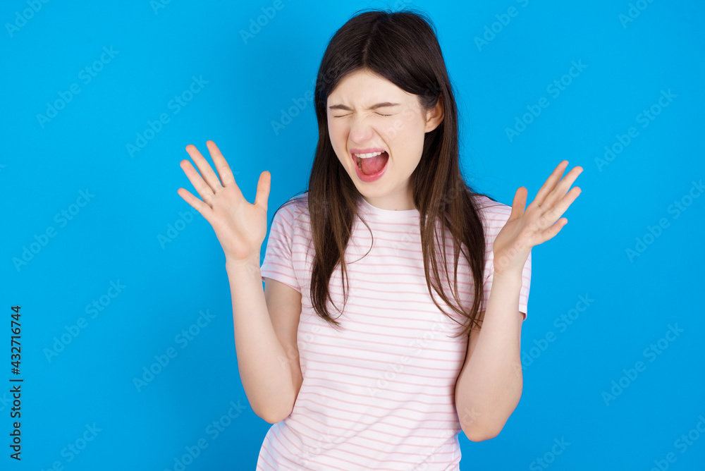 Crazy outraged young beautiful Caucasian woman wearing stripped T-shirt over blue wall screams loudly and gestures angrily yells furiously. Negative human emotions feelings concept Crazy outraged young beautiful Caucasian woman wearing stripped T-shirt over blue wall screams loudly and gestures angrily yells furiously. Negative human emotions feelings concept