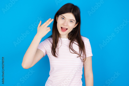young beautiful Caucasian woman wearing stripped T-shirt over blue wall foolishes around shoots in temple with fingers makes suicide gesture. Funny model makes finger gun pistol