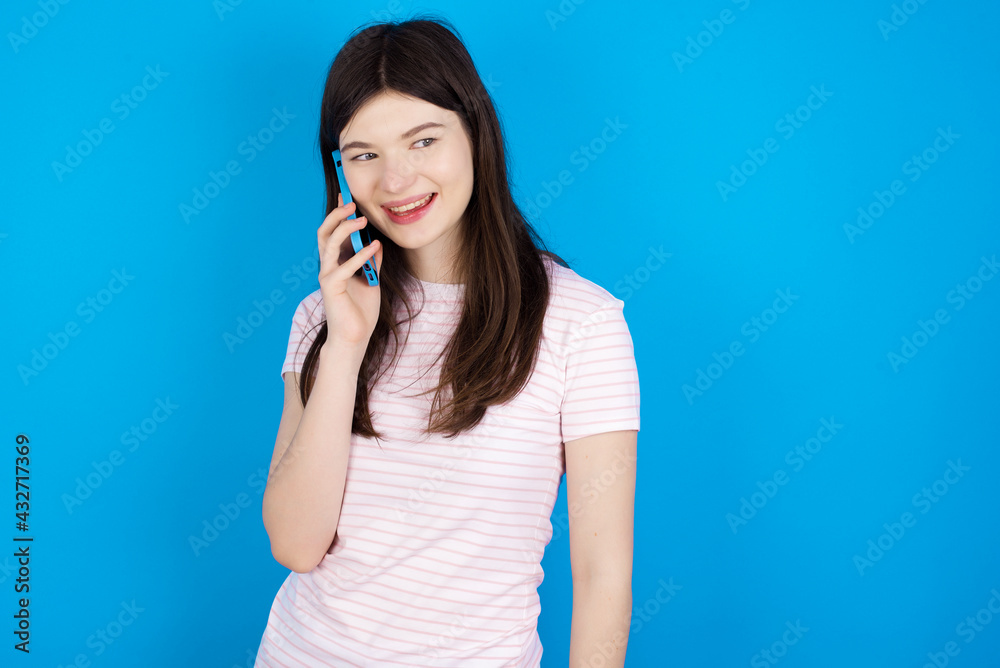 Pleasant looking happy young beautiful Caucasian woman wearing stripped T-shirt has nice telephone conversation and looks aside, has nice mood and smiles positively while talks via cell phone Pleasant looking happy young beautiful Caucasian woman wearing stripped T-shirt has nice telephone conversation and looks aside, has nice mood and smiles positively while talks via cell phone