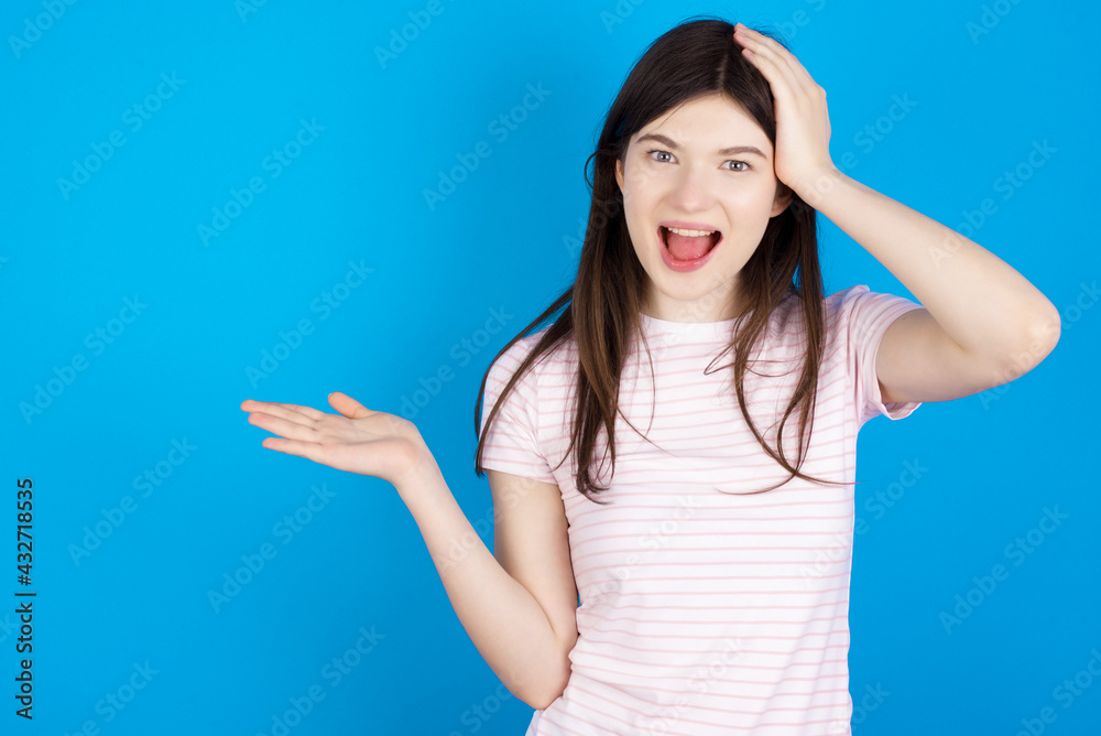 Fototapeta premium Shocked amazed surprised young beautiful Caucasian woman wearing stripped T-shirt over blue wall hold hand offering proposition