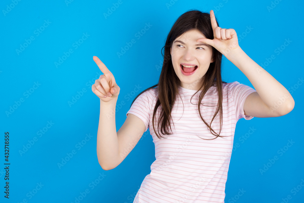 young beautiful Caucasian woman wearing stripped T-shirt over blue wall showing loser sign and pointing at empty space