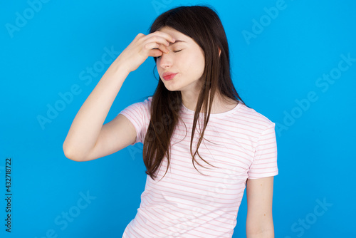 Sad young beautiful Caucasian woman wearing stripped T-shirt over blue wall suffering from headache holding hand on her face