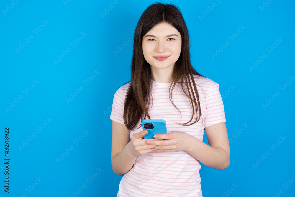 Portrait of serious confident young beautiful Caucasian woman wearing stripped T-shirt over blue wall holding phone in two hands Portrait of serious confident young beautiful Caucasian woman wearing stripped T-shirt over blue wall holding phone in two hands