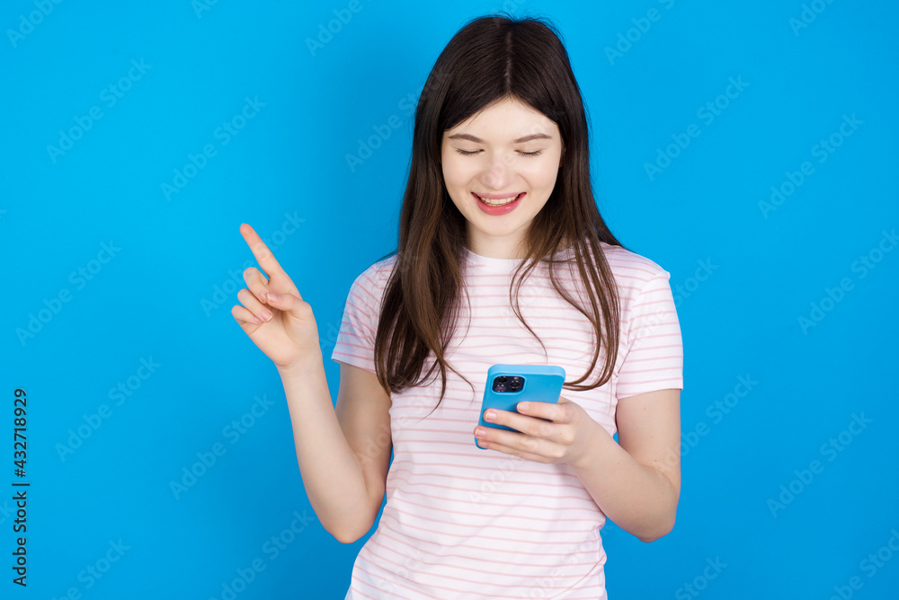 Smiling young beautiful Caucasian woman wearing stripped T-shirt over blue wall pointing finger at blank space holding phone in one hand Smiling young beautiful Caucasian woman wearing stripped T-shirt over blue wall pointing finger at blank space holding phone in one hand