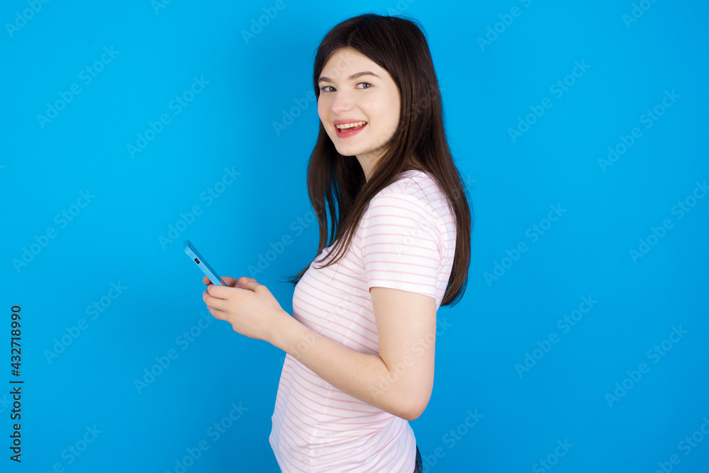 Rear view photo portrait of young beautiful Caucasian woman wearing stripped T-shirt over blue wall using smartphone smiling Rear view photo portrait of young beautiful Caucasian woman wearing stripped T-shirt over blue wall using smartphone smiling