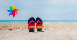 a windmill with rainbow colors next to flip flops on the sand of the beach. the background is turquoise blue and is selectively out of focus. lgbt summer vacation concept. tolerance and respect.