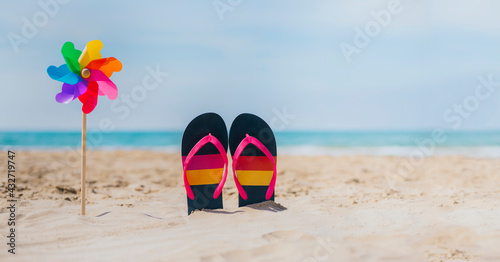 a windmill with rainbow colors next to flip flops on the sand of the beach. the background is turquoise blue and is selectively out of focus. lgbt summer vacation concept. tolerance and respect.