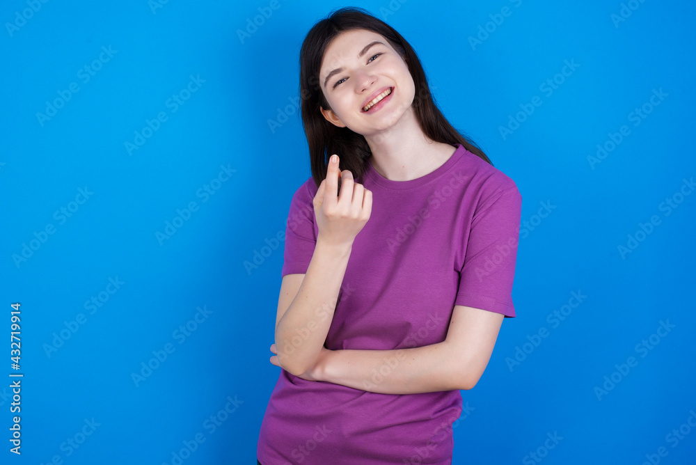 young beautiful Caucasian woman wearing purple T-shirt over blue wall Beckoning come here gesture with hand inviting welcoming happy and smiling young beautiful Caucasian woman wearing purple T-shirt over blue wall Beckoning come here gesture with hand inviting welcoming happy and smiling