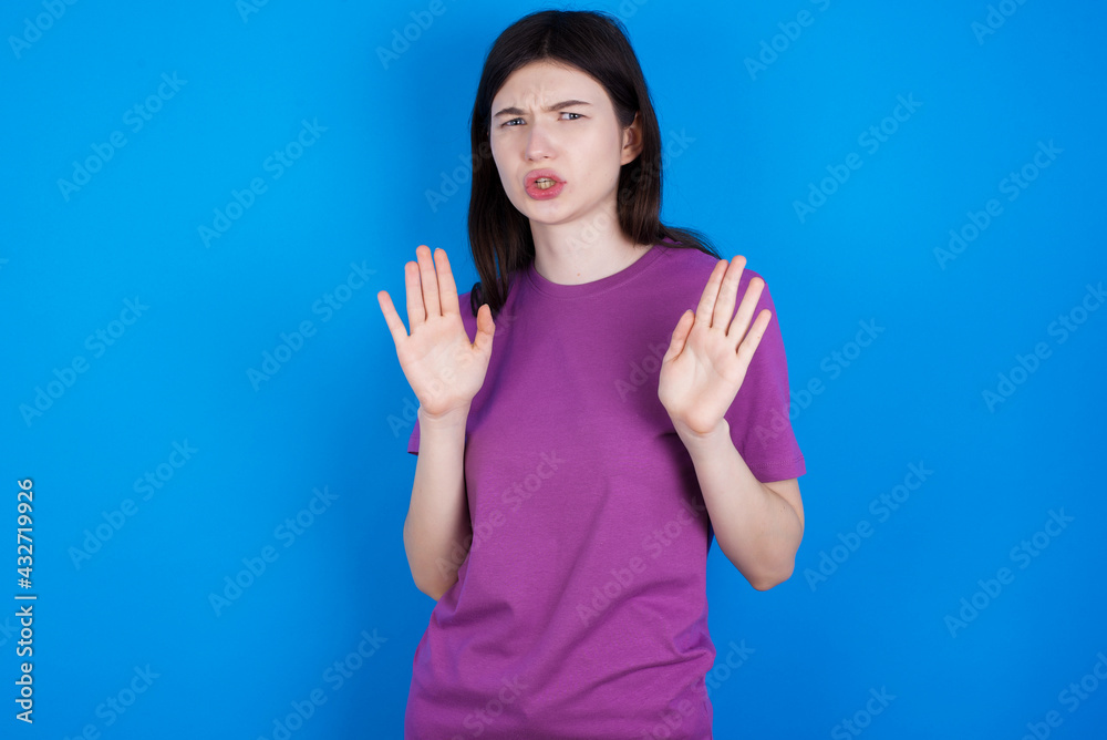 Fototapeta premium young beautiful Caucasian woman wearing purple T-shirt over blue wall Moving away hands palms showing refusal and denial with afraid and disgusting expression. Stop and forbidden.