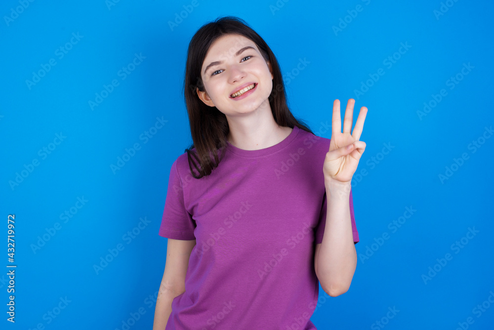 young beautiful Caucasian woman wearing purple T-shirt over blue wall showing and pointing up with fingers number three while smiling confident and happy. young beautiful Caucasian woman wearing purple T-shirt over blue wall showing and pointing up with fingers number three while smiling confident and happy.