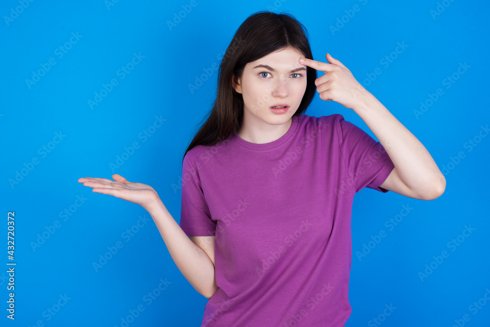 young beautiful Caucasian woman wearing purple T-shirt over blue wall confused and annoyed with open palm showing copy space and pointing finger to forehead. Think about it.