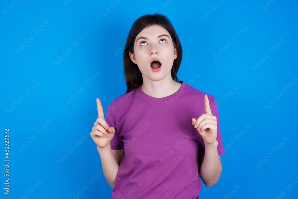 young beautiful Caucasian woman wearing purple T-shirt over blue wall amazed and surprised looking up and pointing with fingers and raised arms. young beautiful Caucasian woman wearing purple T-shirt over blue wall amazed and surprised looking up and pointing with fingers and raised arms.